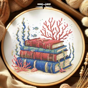 May include: A cross-stitch embroidery in a wooden hoop depicts a stack of books underwater. The books are red, blue, and teal, with coral, seaweed, and fish. A starfish rests on top of the books. The background is white.
