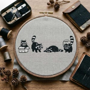 May include: A cross-stitch embroidery hoop featuring four black and white raccoons in various poses. The raccoons are stitched on a light gray fabric, set in a wooden hoop. The scene includes a raccoon in a box, standing, lying down, and holding a jar.