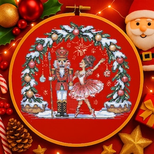 Könnte beinhalten: Ein Kreuzstich-Stickrahmen mit einer Weihnachtsszene mit einem Nussknacker und einer Ballerina. Das Design ist vor einem roten Hintergrund platziert und von einem Holzrahmen umgeben. Ornamente und festliches Grünzeug vervollständigen das Weihnachtsthema.