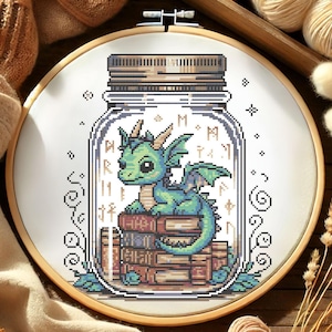 May include: Cross-stitch embroidery in a wooden hoop. The design features a green dragon sitting on a stack of books inside a mason jar. The jar has a brown lid and is surrounded by decorative swirls and stars.