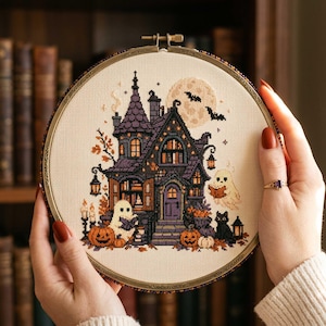 May include: A circular embroidery hoop containing a cross-stitch pattern of a purple haunted house with orange pumpkins, two white ghosts, a black cat, and a full moon with bats against a cream-colored fabric background.