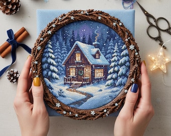 May include: A cross-stitch embroidery of a snow-covered cabin in a winter forest, framed in a brown wreath with snowflake accents. The embroidery is held by hands with painted nails. Cinnamon sticks, pine cones, and scissors are also visible.