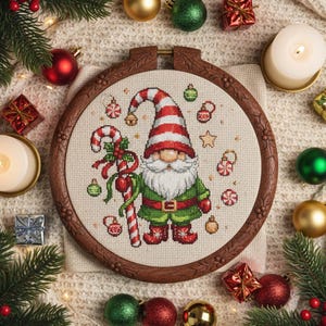 Op de afbeelding: Kruissteekborduurwerk van een kerstkabouter in een houten hoepel. De kabouter draagt een rood-witte gestreepte hoed en een groene outfit, met een zuurstok. Ornamenten en feestelijke decoraties omringen het borduurwerk.