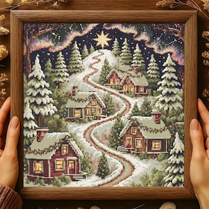Puede incluir: Un bordado enmarcado que representa una escena de un pueblo invernal. La obra presenta casas cubiertas de nieve a lo largo de un camino sinuoso, rodeadas de árboles de hoja perenne. Una gran estrella brilla en el cielo nocturno sobre el pueblo.