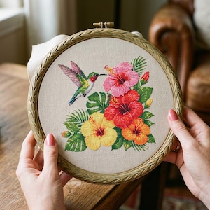 Può includere: Ricamo a punto croce di un colibrì e fiori di ibisco in un telaio di legno. Il colibrì ha piume verdi, rosse e blu. I fiori sono rosa, rossi, gialli e arancioni. Il ricamo è tenuto in mano.