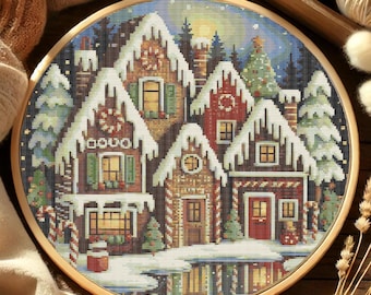 May include: A cross-stitch embroidery of a winter village scene, featuring snow-covered houses with red and brown facades. The houses have lit windows and are surrounded by snow-covered trees. The embroidery is set in a wooden hoop.