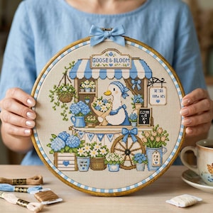 May include: A circular embroidery hoop containing a cross-stitch pattern of a goose in a flower shop. The shop features a blue and white striped awning with the text GOOSE & BLOOM, blue hydrangeas, potted plants, and a wooden wagon wheel.