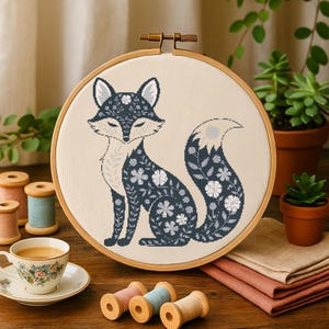 May include: A black and white embroidery hoop with a floral pattern fox design. The fox is sitting with its tail curled around its body. The hoop is on a wooden table with other embroidery supplies.