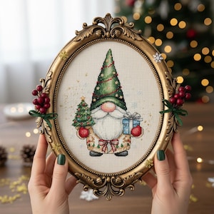 Könnte beinhalten: Ein ovaler, goldfarbener Rahmen hält ein Kreuzstichdesign eines Weihnachtszwergs. Der Zwerg trägt einen grünen Hut und hält einen Weihnachtsbaum, ein Geschenk und Ornamente. Der Rahmen ist mit roten Beeren und grünen Bändern verziert.