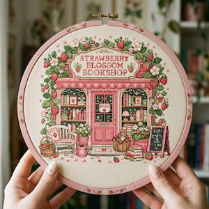 Op de afbeelding: Een ronde borduurring met een kruissteekontwerp van een winkel genaamd STRAWBERRY BLOSSOM BOOKSHOP. De scène toont een roze winkelpui versierd met aardbeienranken, bloemen, boekenplanken, een bankje en een bordje met de tekst OPEN.