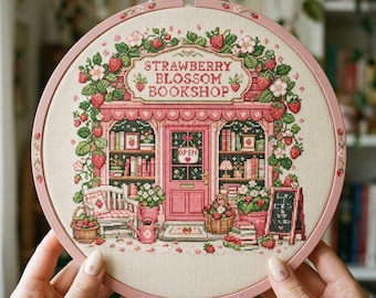 May include: A circular embroidery hoop containing a cross-stitch design of a shop titled STRAWBERRY BLOSSOM BOOKSHOP. The scene features a pink storefront adorned with strawberry vines, flowers, bookshelves, a bench, and a sign reading OPEN.
