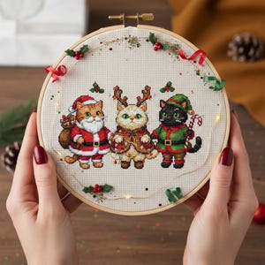 May include: A cross-stitch embroidery hoop featuring three cats dressed in Christmas attire. The cats are in Santa, reindeer, and elf costumes. The embroidery is decorated with red ribbons, holly, and small lights, set in a wooden hoop.