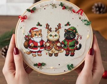 May include: A cross-stitch embroidery hoop featuring three cats dressed in Christmas attire. The cats are in Santa, reindeer, and elf costumes. The embroidery is decorated with red ribbons, holly, and small lights, set in a wooden hoop.