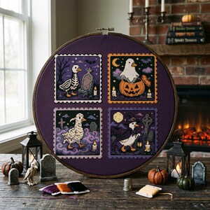 May include: A circular wooden embroidery hoop containing a dark purple fabric with four square cross-stitch designs. Each square features a white duck in a Halloween setting, including a skeleton duck, a ghost in a pumpkin, a duck in fog, and a duck in a witch hat holding a lantern.