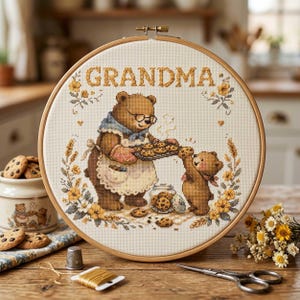 May include: A cross-stitch embroidery hoop featuring a brown bear wearing glasses and an apron, baking cookies with a smaller bear. The word "GRANDMA" is stitched across the top. The design includes yellow flowers and wheat stalks.