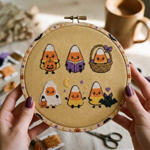 May include: A circular wooden embroidery hoop containing a tan fabric with six cross-stitched candy corn characters. The designs include a candy corn holding a pumpkin, reading a book, sitting in a basket, wearing a ghost costume, standing with a crescent moon, and next to a bat.