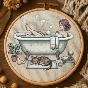 Puede incluir: Bordado de punto de cruz en un aro de madera que representa a una mujer en un baño de burbujas. Un gato atigrado descansa sobre una alfombra cerca de la bañera. Una pequeña mesa contiene una copa de vino y un libro. También hay una planta en maceta y velas.