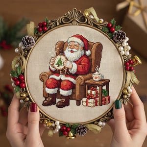 Può includere: Un ricamo a punto croce incorniciato di Babbo Natale su una sedia, che tiene in mano un biscotto con un disegno di albero di Natale. La cornice dorata ha elementi decorativi, tra cui agrifoglio, bacche e nastri. Un tavolino con regali e una tazza di caffè è accanto a Babbo Natale.