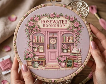 May include: A circular wooden embroidery hoop containing a detailed cross-stitch design of a pink storefront labeled Rosewater Bookshop. The scene features shelves of books, climbing roses, a watering can, and a sign that reads Books & Beautiful Things.