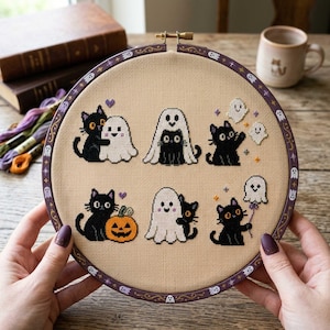 Peut inclure: Un tambour à broder rond contenant un tissu beige avec des motifs au point de croix de chats noirs et de fantômes blancs. Les motifs incluent des chats embrassant des fantômes, un chat sous un drap de fantôme, un chat avec une citrouille.