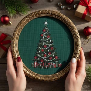Op de afbeelding: Een ingelijst kruissteekontwerp van een kerstboom. De boom is groen met rode ballen en witte guirlande. De ronde lijst is goudkleurig. De achtergrond is donkergroene stof. Kerstversieringen omringen de afbeelding.