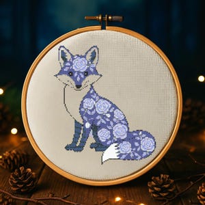 Puede incluir: Un aro de bordado de punto de cruz con un diseño de zorro morado. El zorro está cubierto de flores moradas y tiene una cola blanca.