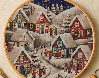 May include: A cross-stitch embroidery of a winter village scene, set within a round wooden hoop. The scene depicts snow-covered houses with lit windows, a winding road, and figures in red coats. The background shows a starry night sky.