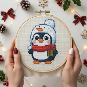 May include: A cross-stitch embroidery of a penguin wearing a white and blue hat and a red scarf. The penguin is holding a green and red gift. The embroidery is in a wooden hoop, perfect for holiday decor.