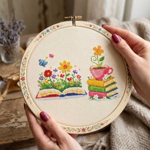 May include: A cross-stitch embroidery hoop featuring a colorful floral design. The design includes an open book overflowing with flowers and a stack of books with a teacup and flower. The hoop is held by hands.