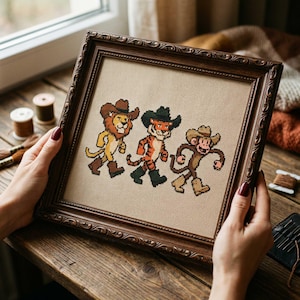 May include: Cross-stitch artwork in a dark brown ornate frame. The artwork depicts a lion, tiger, and monkey, each wearing a cowboy hat and boots. The animals are stitched in shades of yellow, orange, brown, and black on a beige background.
