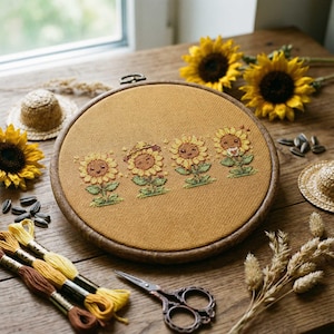 May include: A cross-stitch embroidery hoop showing four smiling sunflowers with hats and accessories. The embroidery is set against a mustard-coloured fabric, framed by a wooden hoop. The scene includes sunflowers, embroidery floss, and a pair of scissors.