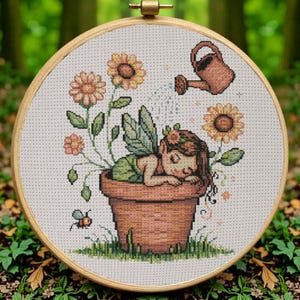 Op de afbeelding: Kruissteekborduurwerk in een houten hoepel. Het ontwerp toont een slapende fee in een bloempot, omringd door zonnebloemen en een gieter. Een kleine bij is ook in het ontwerp opgenomen. Het kleurenpalet omvat tinten groen, bruin en geel.