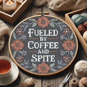 Puede incluir: Un bastidor de bordado de punto de cruz con el texto "Fueled by Coffee and Spite" en blanco. El diseño presenta acentos florales en melocotón y verde azulado sobre un fondo gris oscuro, en un bastidor de madera. Una taza de té y lana están cerca.