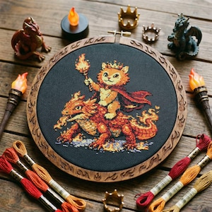 May include: A cross-stitch embroidery featuring a cartoon cat wearing a crown and cape, riding a dragon, holding a torch. The embroidery is set in a wooden hoop with a decorative border. Additional dragon figurines, crowns, and torch-shaped lights surround the embroidery.