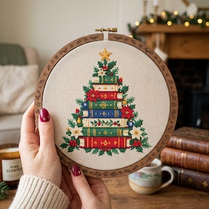 May include: A circular wooden embroidery hoop containing a cross-stitch design of a Christmas tree formed by a stack of colourful books. The tree is decorated with red poinsettias, green holly leaves, and a yellow star at the very top.
