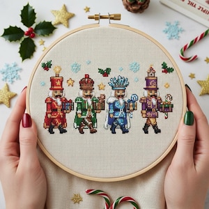 Puede incluir: Un bastidor de bordado de punto de cruz con cuatro figuras de cascanueces coloridos. Cada cascanueces viste un uniforme diferente, sosteniendo regalos y linternas. El bordado está sobre una tela color crema, en un aro de madera.
