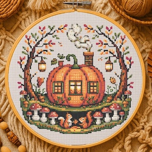 May include: A cross-stitch embroidery in a wooden hoop features a pumpkin house with a chimney, windows, and a green stem. Squirrels sit below the pumpkin, surrounded by mushrooms and trees with autumn leaves. Two lanterns hang from the trees. The color palette includes orange, green, brown, and white.