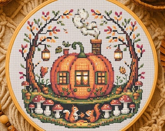 May include: A cross-stitch embroidery in a wooden hoop features a pumpkin house with a chimney, windows, and a green stem. Squirrels sit below the pumpkin, surrounded by mushrooms and trees with autumn leaves. Two lanterns hang from the trees. The color palette includes orange, green, brown, and white.