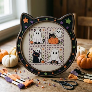 Peut inclure: Un cadre en bois noir en forme de chat contenant une broderie au point de croix avec quatre panneaux carrés représentant des chats noirs et des fantômes blancs. Le cadre est orné de petites étoiles orange, violettes et vertes sur fond beige.