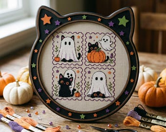 May include: A black cat-shaped wooden frame containing a cross-stitch sampler with four square panels featuring black cats and white ghosts. The frame is decorated with small orange, purple, and green stars. The sampler is on light beige fabric.