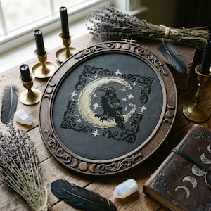 Könnte beinhalten: Ein gerahmtes Kreuzstichbild mit einem schwarzen Raben auf einem Mond, umgeben von Sternen, auf dunklem Hintergrund. Der Holzrahmen hat ein dekoratives Design. Die Szene befindet sich auf einem Holztisch mit Kerzen, Federn und getrocknetem Lavendel.