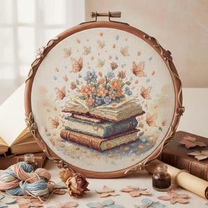May include: A cross-stitch embroidery hoop featuring a stack of books with flowers and butterflies. The design is in shades of beige, brown, blue, and peach. The wooden hoop has decorative metal accents and is surrounded by craft supplies.