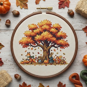 May include: A cross-stitch embroidery in a wooden hoop depicts an autumn scene. A large tree with orange and yellow leaves is the focal point, with woodland creatures gathered beneath. Pumpkins, acorns, and fall leaves surround the hoop.