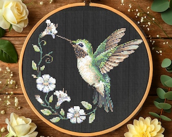 May include: Cross-stitch embroidery of a hummingbird with green, brown, and white details, hovering near white flowers. The design is set against a black background within a wooden embroidery hoop. The image is surrounded by flowers and greenery.