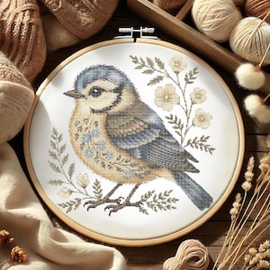 Peut inclure: Un oiseau bleu avec des marques brunes et blanches est assis sur une branche avec des fleurs blanches. L'oiseau est entouré de feuilles vertes et est brodé sur une toile blanche.