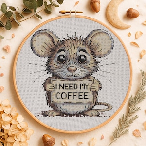 Peut inclure: Broderie au point de croix d'une souris tenant une pancarte avec l'inscription "I NEED MY COFFEE". La souris est marron et beige avec de grands yeux et des moustaches. La broderie est dans un cercle en bois, sur fond neutre.
