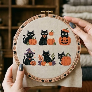May include: A circular wooden embroidery hoop containing a beige fabric panel with six cross-stitch designs of black cats and orange pumpkins. The hoop frame is decorated with small engraved images of cats, pumpkins, and autumn leaves.