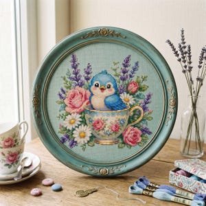 Puede incluir: Un bordado de punto de cruz enmarcado con un pájaro azul en una taza floral, rodeado de rosas rosas, lavanda y margaritas. El marco redondo está pintado de color turquesa claro con detalles decorativos. Una encantadora pieza de decoración del hogar.