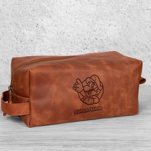 May include: A brown leather toiletry bag with a zipper closure and a handle on the side. The bag features an engraved image of Super Mario with the text "SUPER MARIO" below the image.