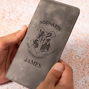 May include: A gray leather wallet featuring the Hogwarts crest and the name "JAMES" engraved on the front. The wallet is held in two hands and appears to be made of a soft, textured material. The design is a nod to the popular fantasy series.